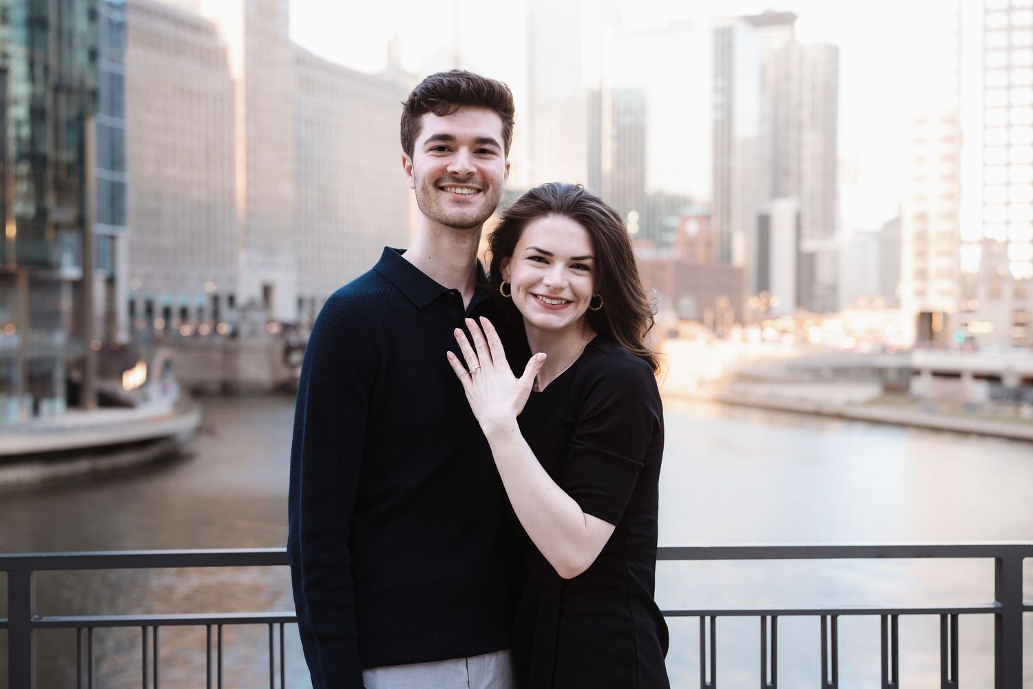 Newly engaged smiling couple posing on bridge, woman showing engagement ring with city river backdrop.
