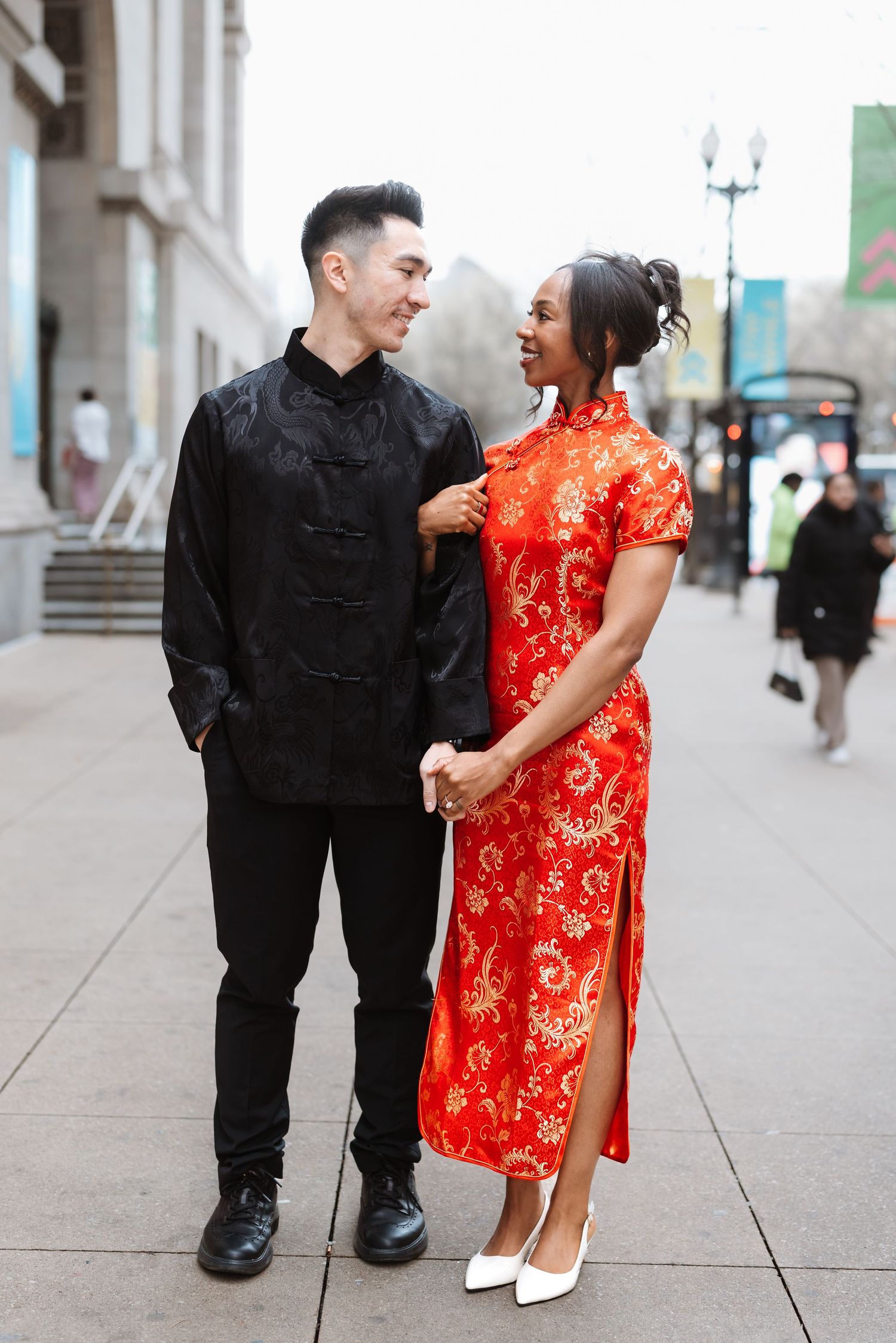 Smiling interracial couple holding hands on city sidewalk wearing traditional Chinese red and black attire.
Chicago Wedding Photographer