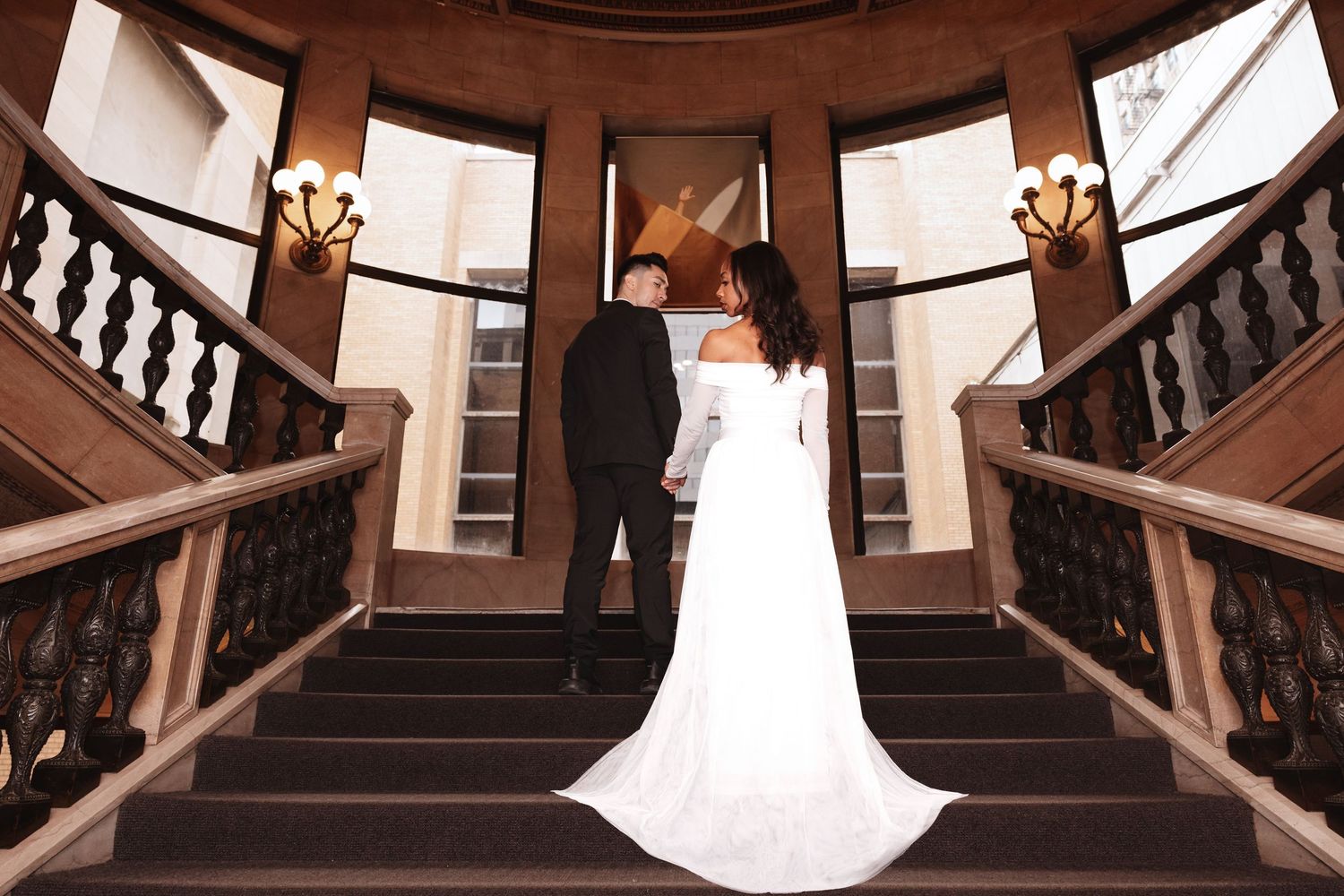 Bride and groom viewed from behind on elegant curved staircase with ornate railings.
