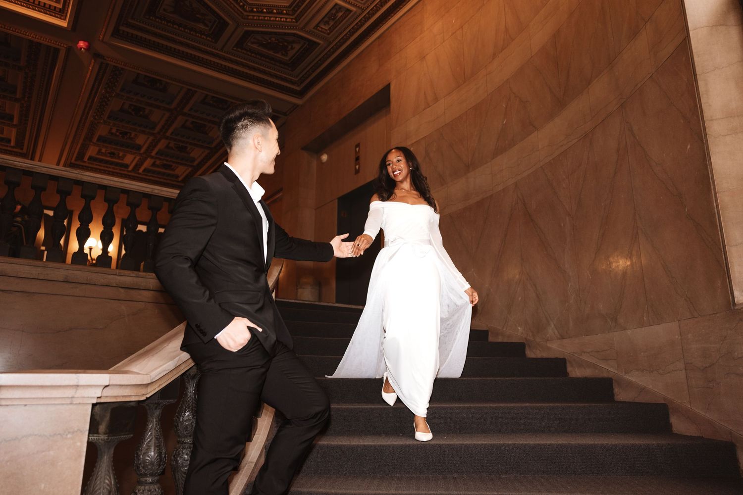 Bride in white jumpsuit laughing as groom leads her up ornate marble staircase.