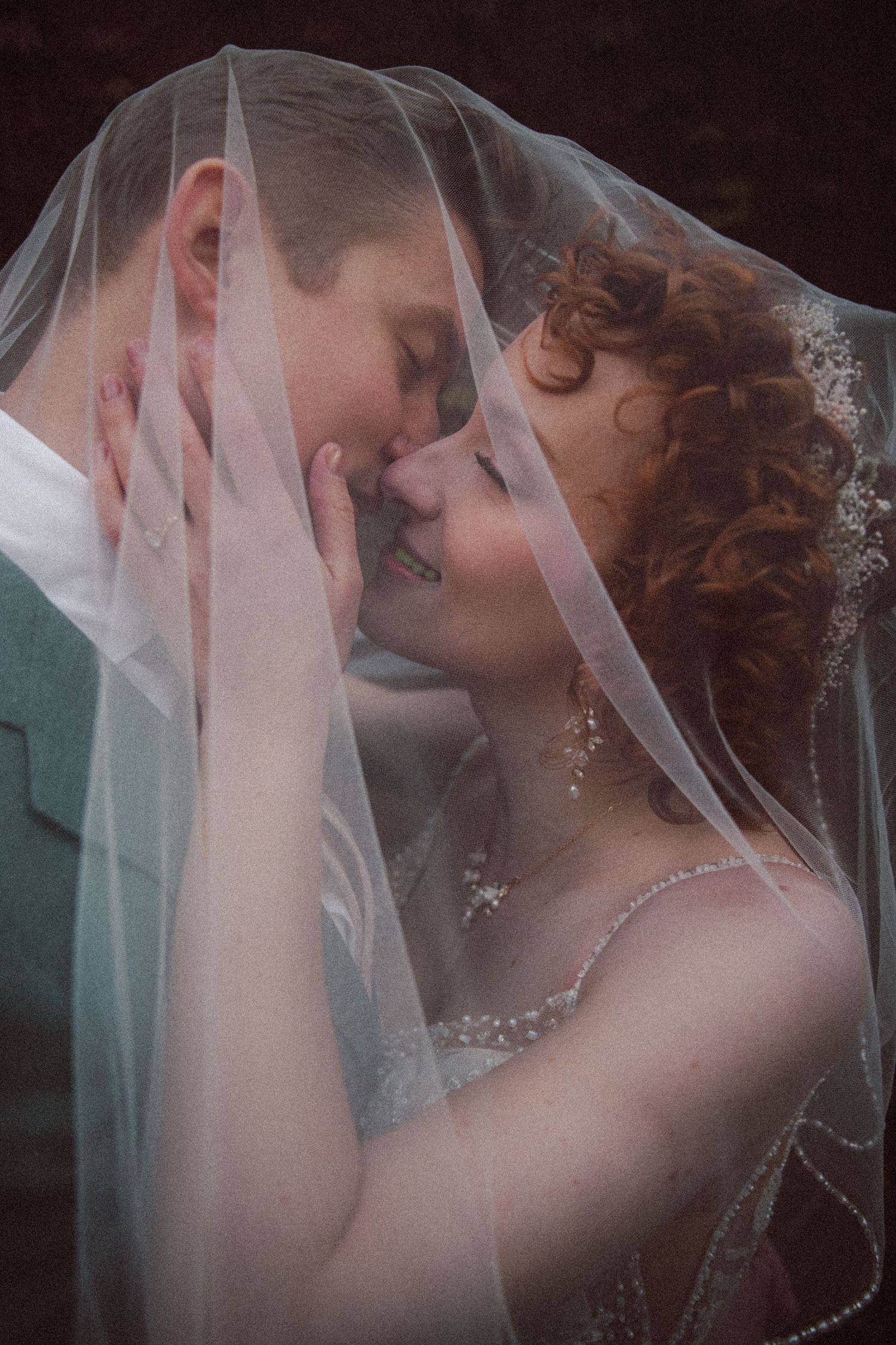Bride with red curly hair and groom share tender moment together beneath her flowing veil.