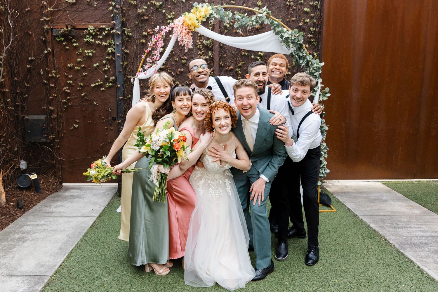 Bride and groom smiling with wedding party including bridesmaids and groomsmen in front of floral arch.
