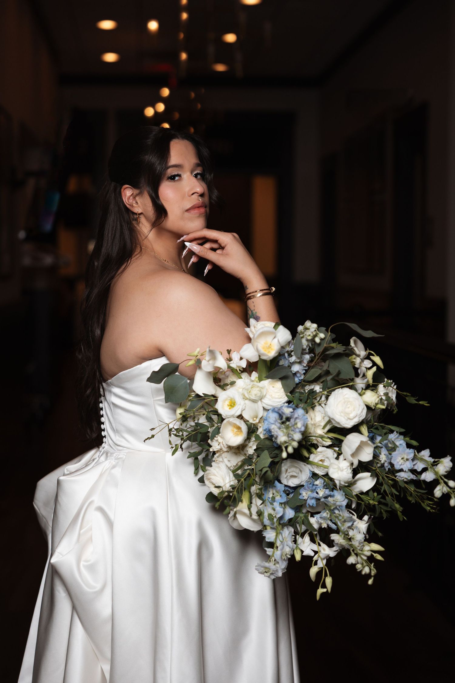 Bride in strapless white satin gown holding blue and white floral bouquet poses elegantly in dimly lit venue.