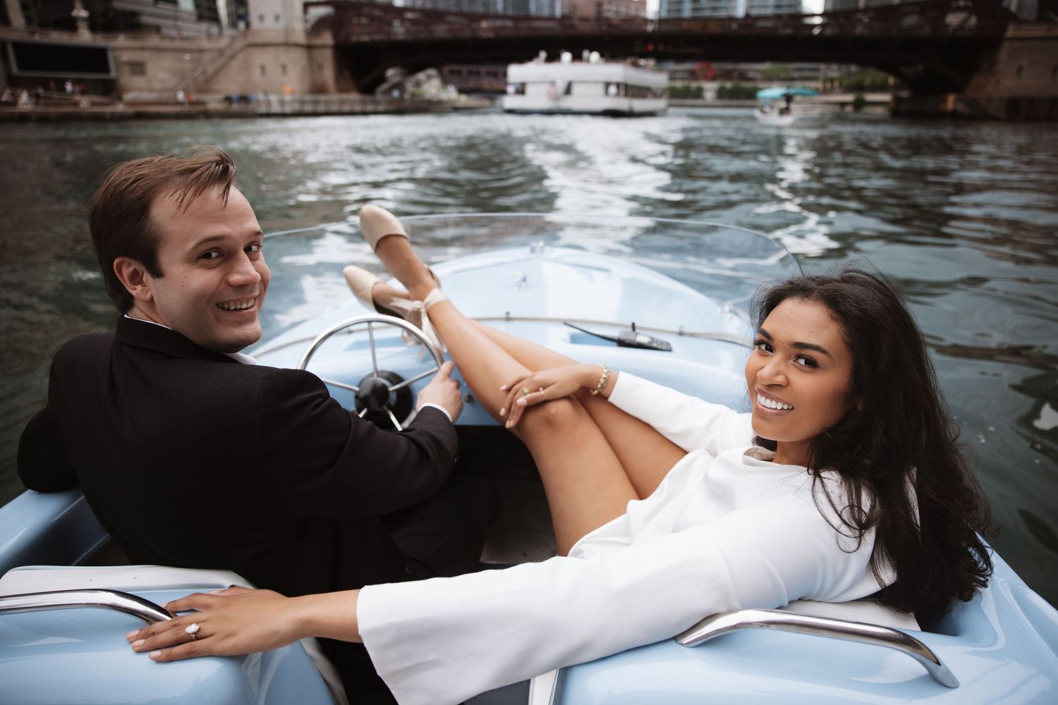 Couple relaxing on a classic boat on the river, woman in white dress smiling, man in black suit, urban waterway backdrop.