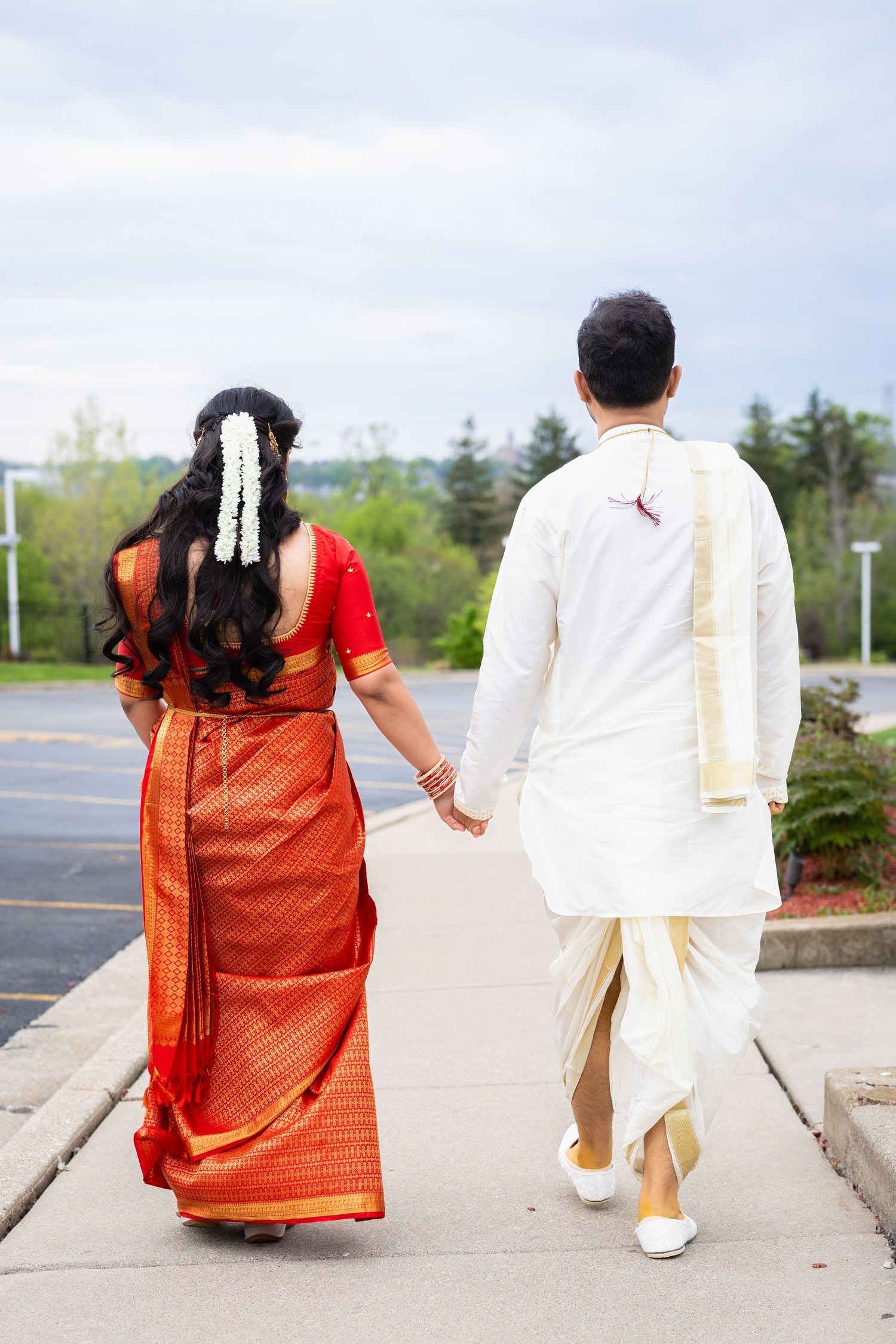 Indian bride and groom walking hand in hand from behind, bride in red saree with floral hair accessory.