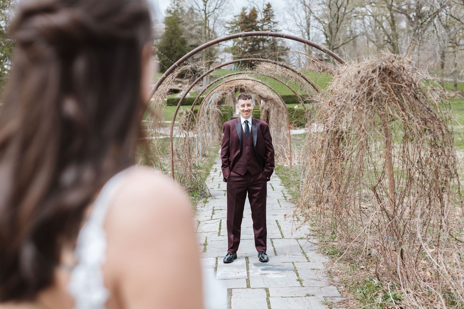 Groom in burgundy suit stands under garden arch, bride in white dress in foreground, outdoor wedding photo.