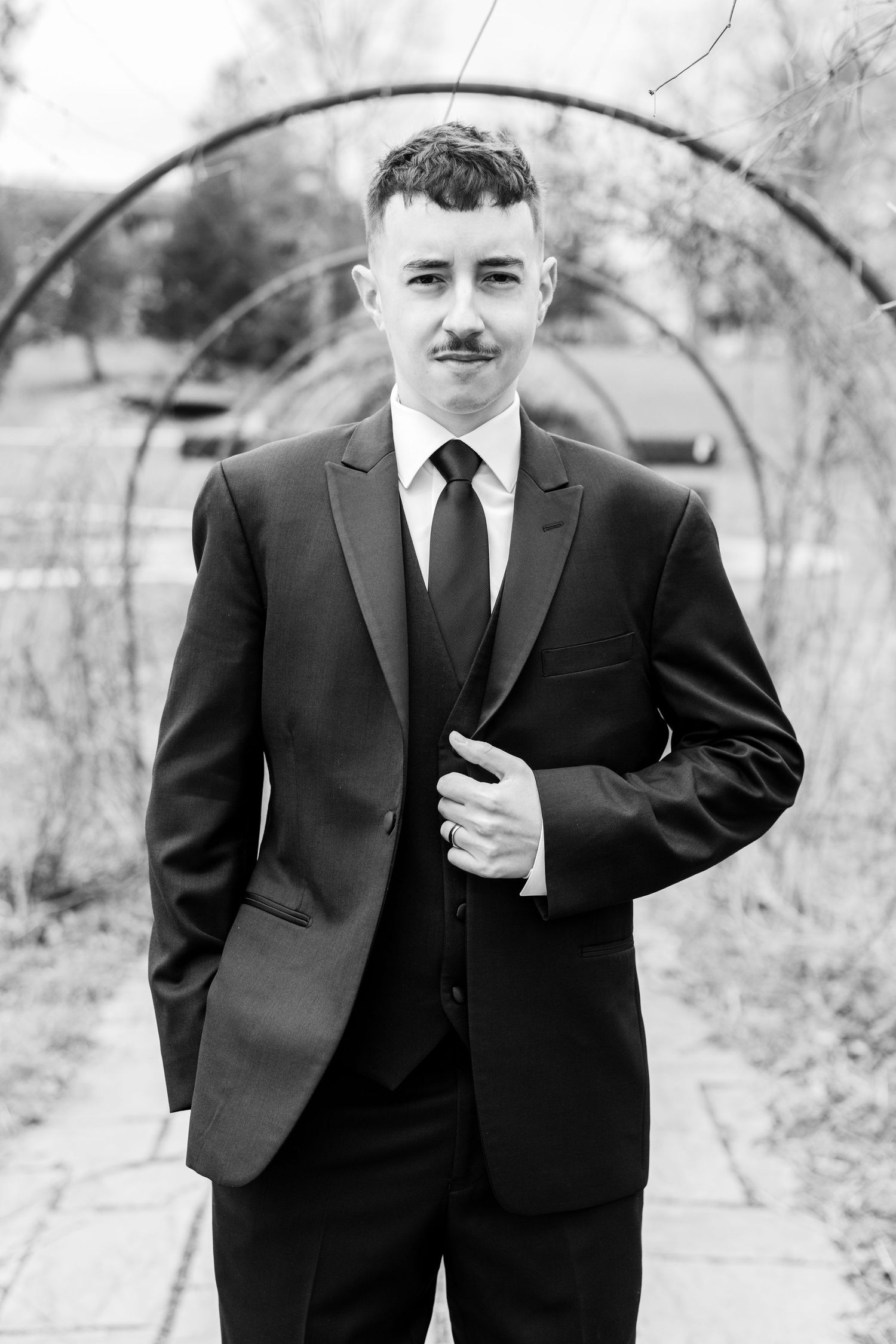 Black and white portrait of a young man in a suit and tie with a mustache, posing outdoors near an arch.