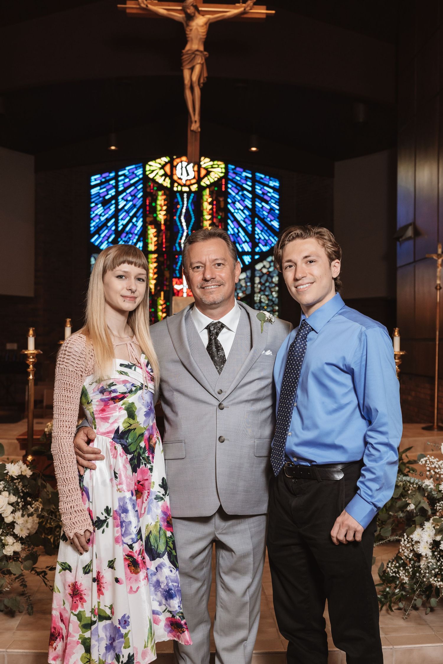 Family portrait of three people posing in front of a crucifix and stained glass window inside a church.