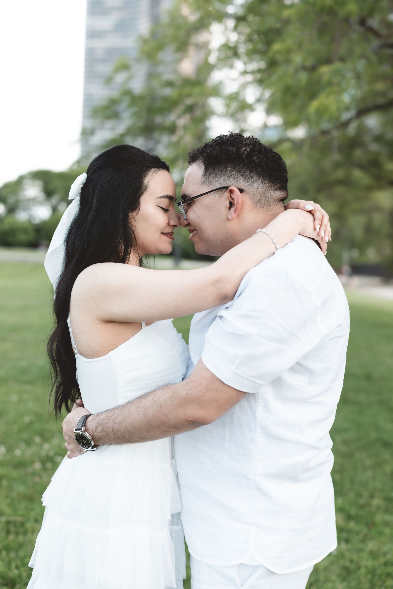 Couple in white outfits sharing a romantic moment in a park, foreheads touching.
