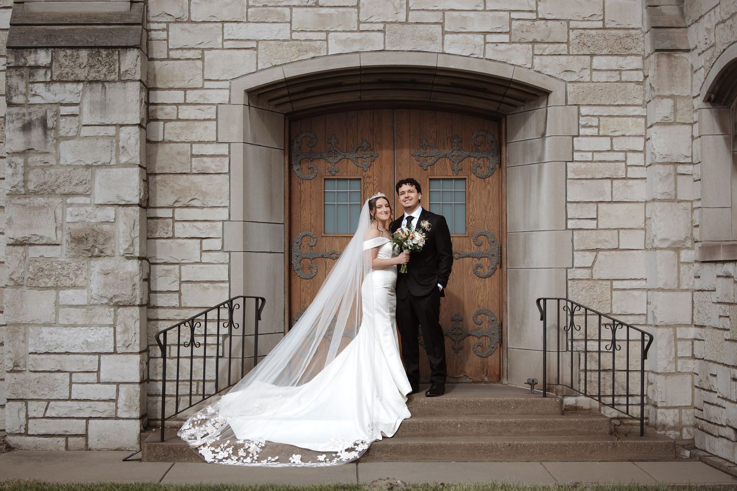 Bride and groom pose on stone church steps in elegant wedding attire with long veil trailing behind.