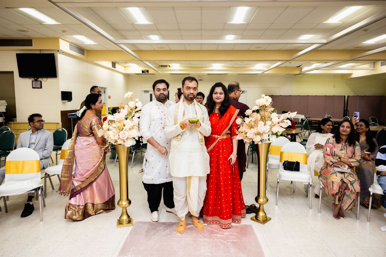 Indian groom in white kurta escorted down aisle by parents during traditional Hindu wedding ceremony.