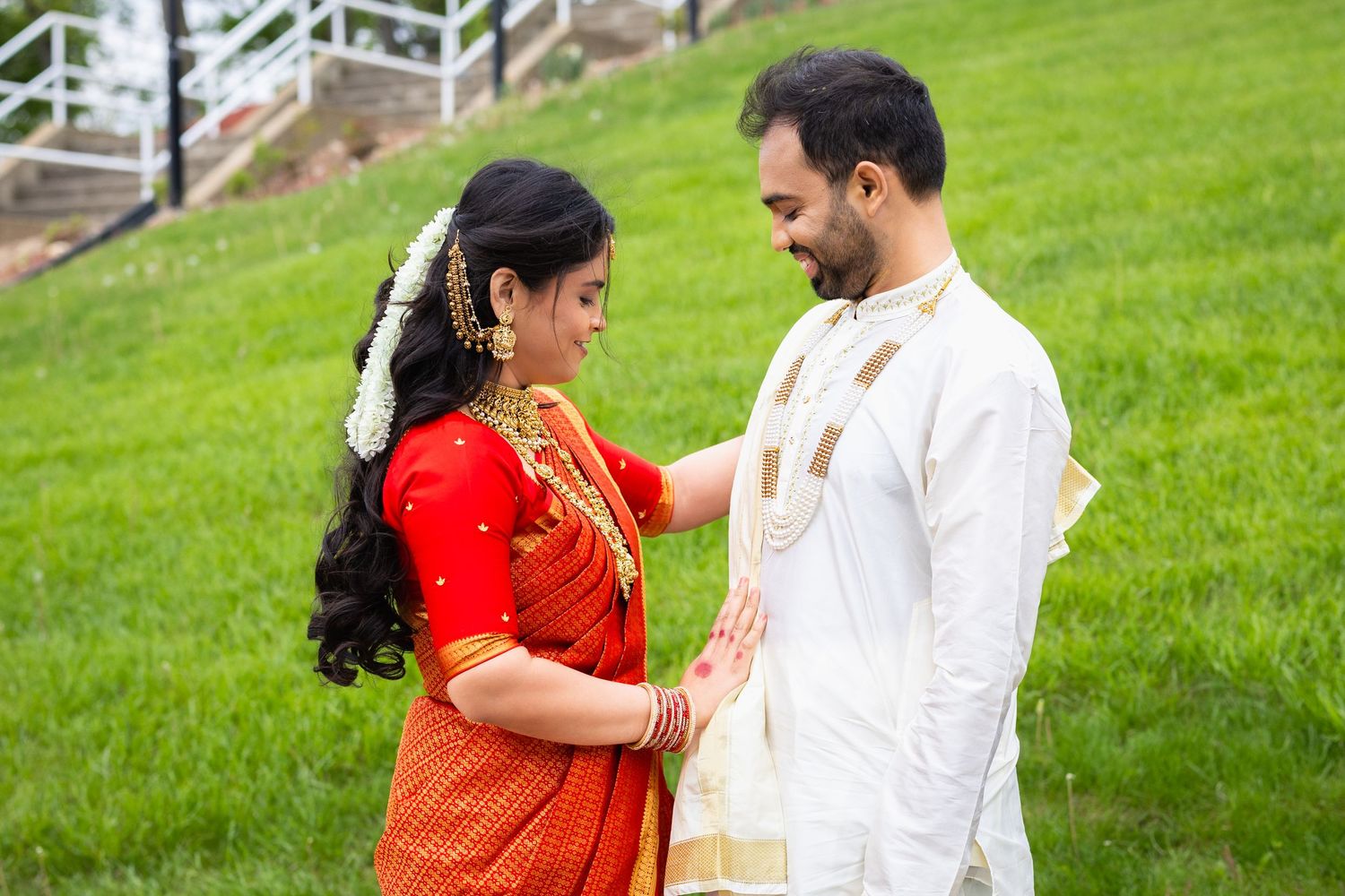 Indian couple in traditional wedding attire, bride in red saree with gold jewelry, groom in white sherwani, standing on green lawn.