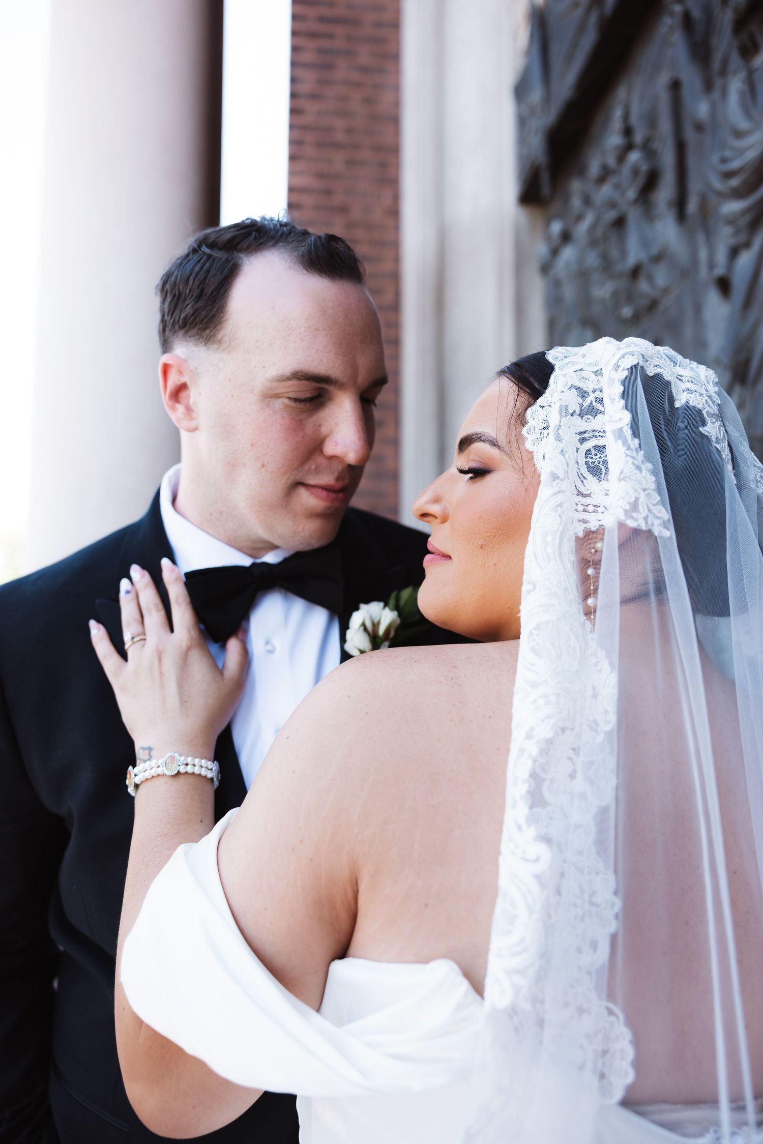 Groom in black bow tie gazing at bride wearing lace mantilla veil against stone church exterior.