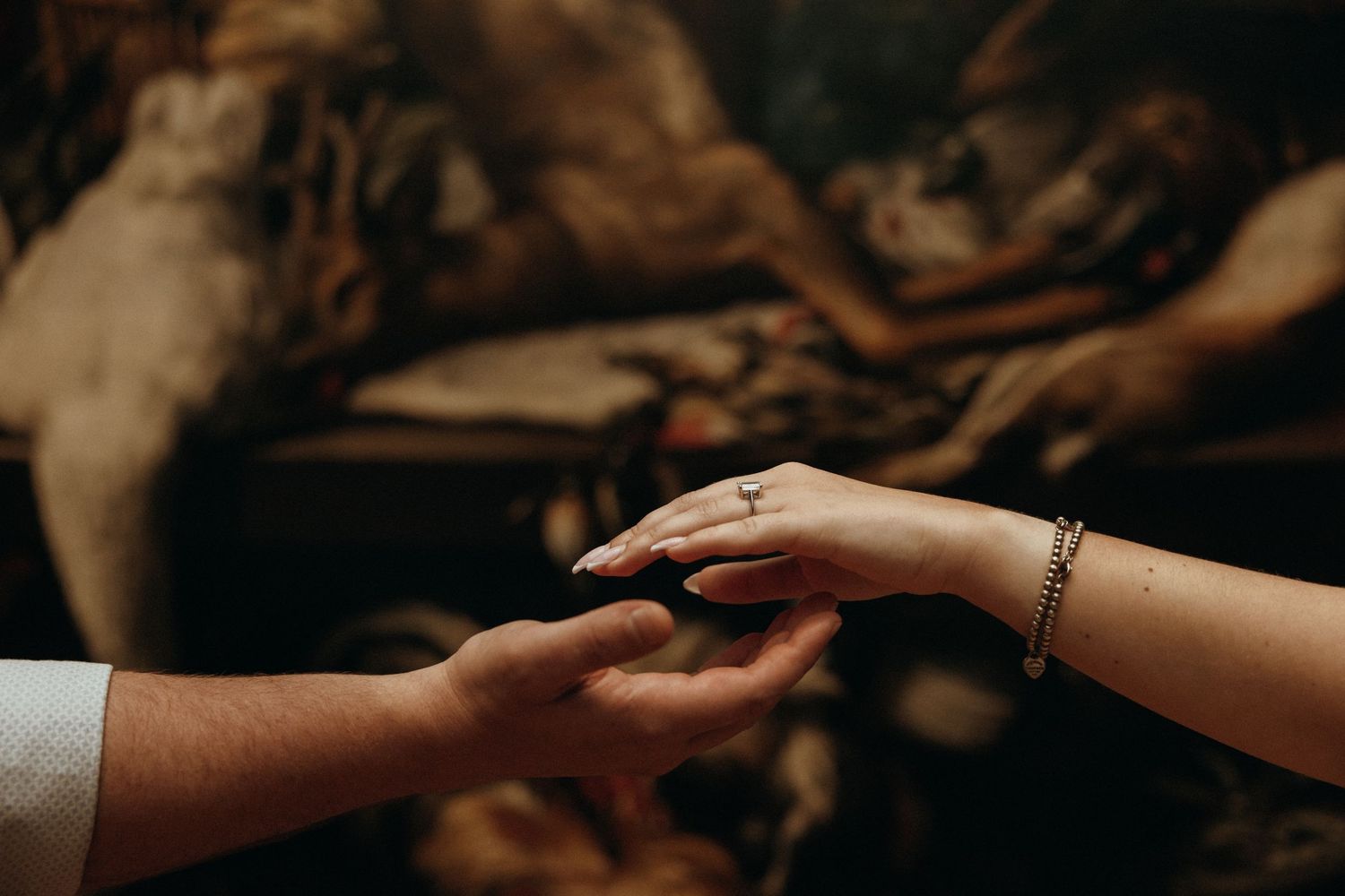 Two hands gently touching, one wearing a delicate cross ring and bracelet, against a dark artistic background.