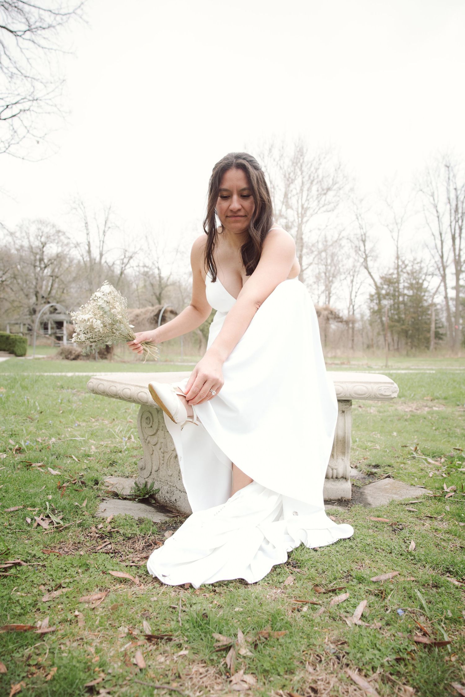 Bride in white wedding dress sitting on rustic stone bench outdoors, holding small white bouquet in a park setting.