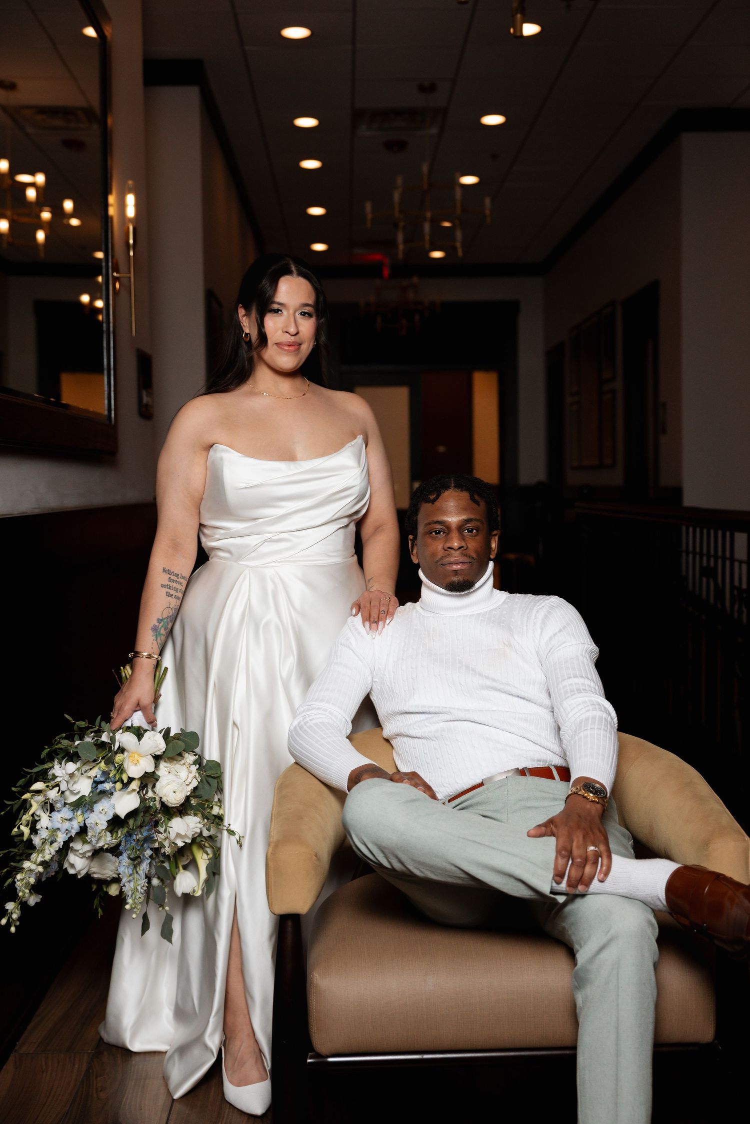 Bride in sleek white satin gown holding bouquet stands beside groom in white turtleneck seated in elegant indoor venue.