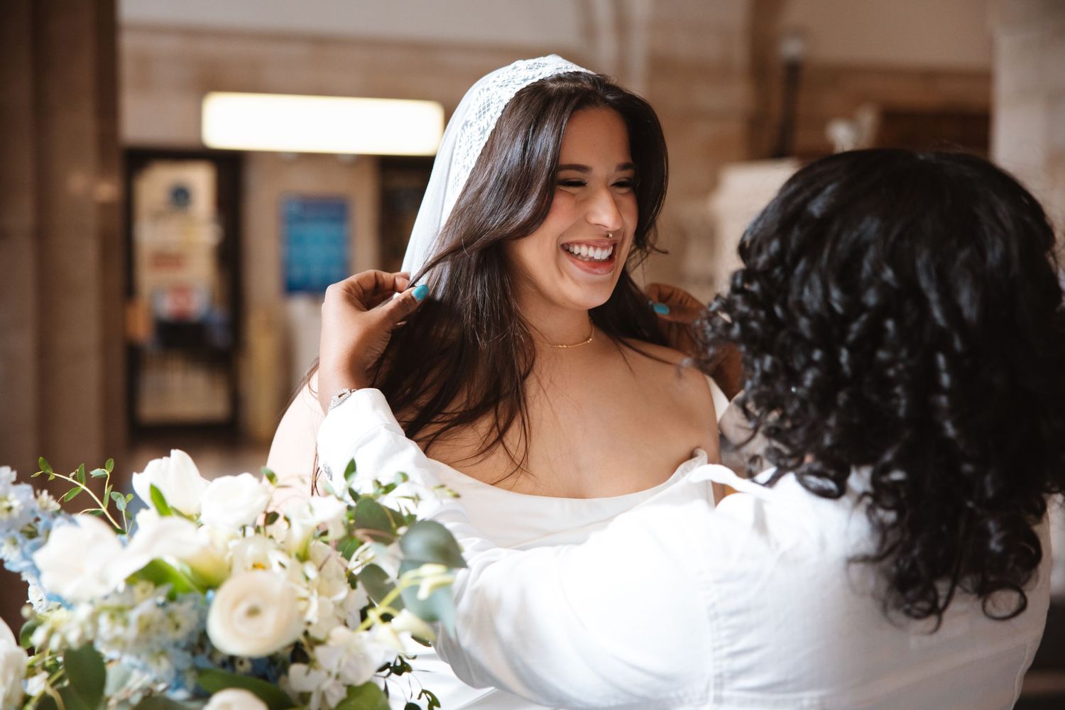 A bride smiles joyfully as a woman adjusts her white veil, with elegant blue and white floral arrangements nearby.