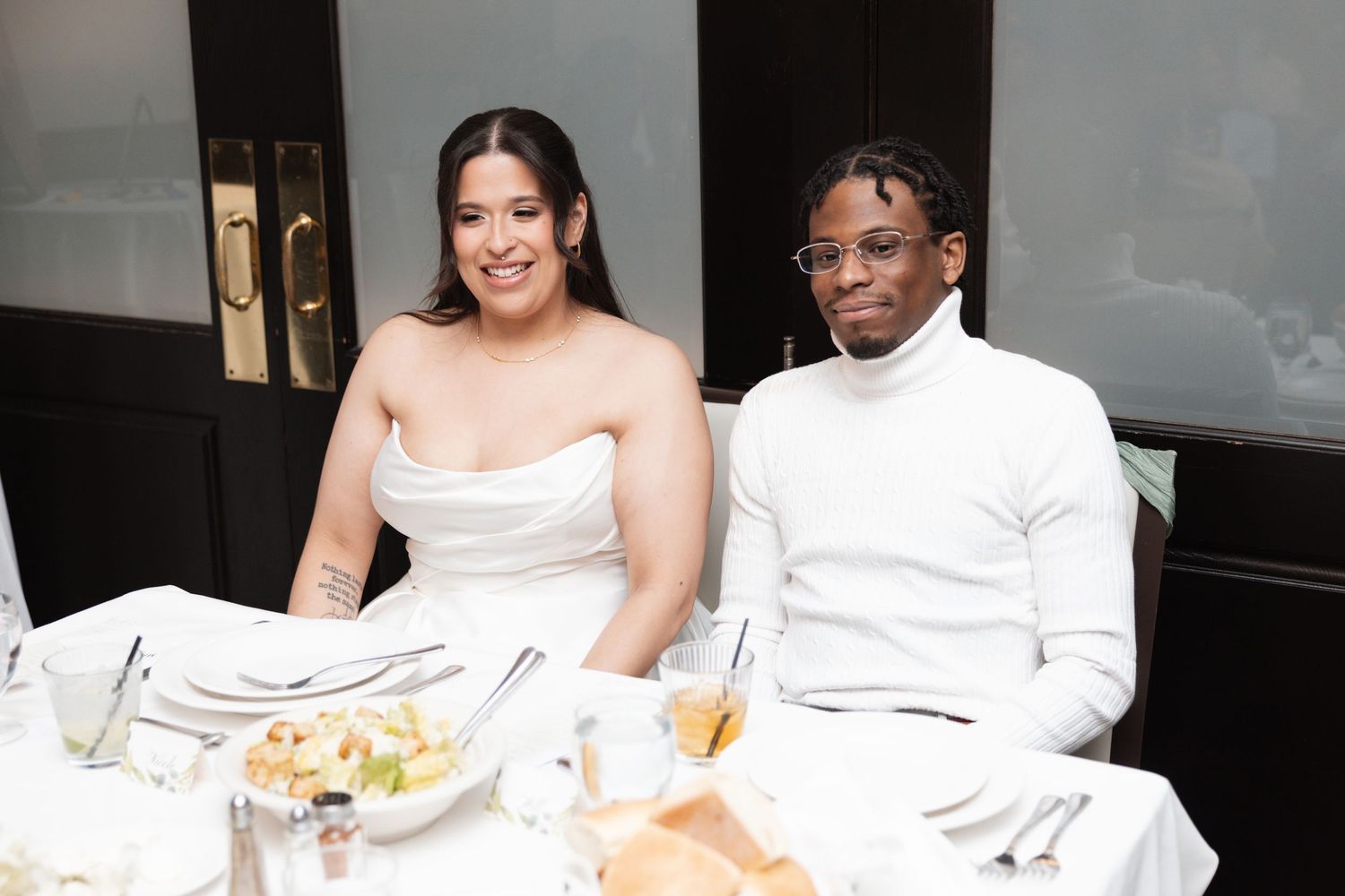 Bride in strapless white gown and groom in white turtleneck sit together at wedding reception table.