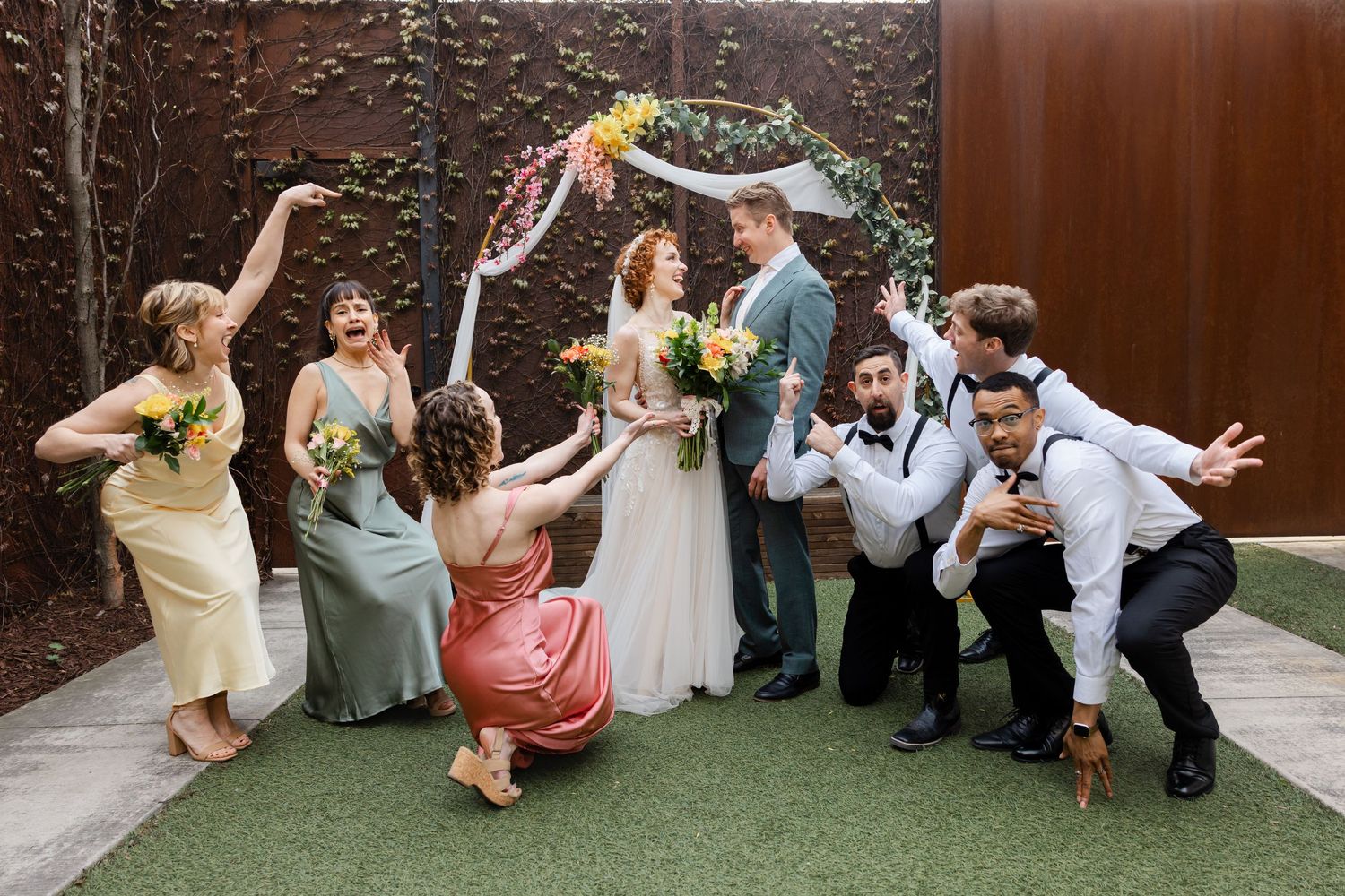 Wedding party celebrates joyfully outdoors under floral arch, bridesmaids in colorful dresses with groomsmen posing playfully on green turf.