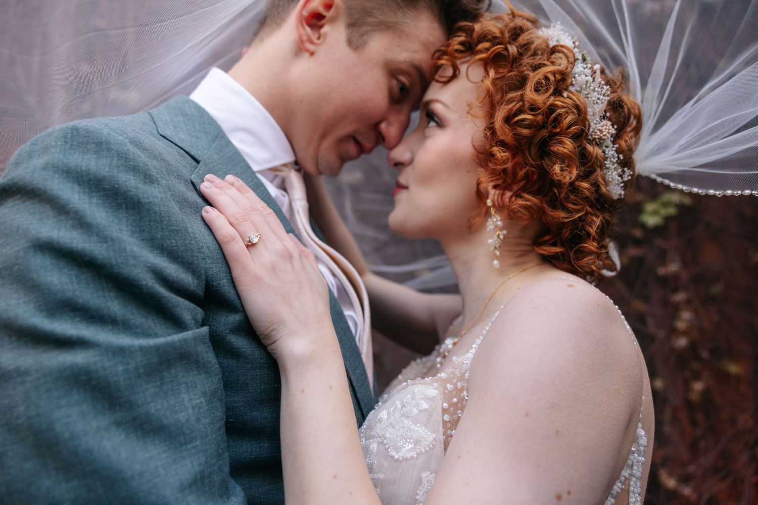 Bride with red curly hair and groom touch foreheads tenderly under a flowing white veil outdoors.