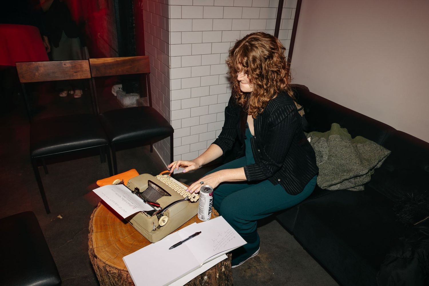 Woman in green dress types on vintage typewriter seated on couch in dimly lit venue.