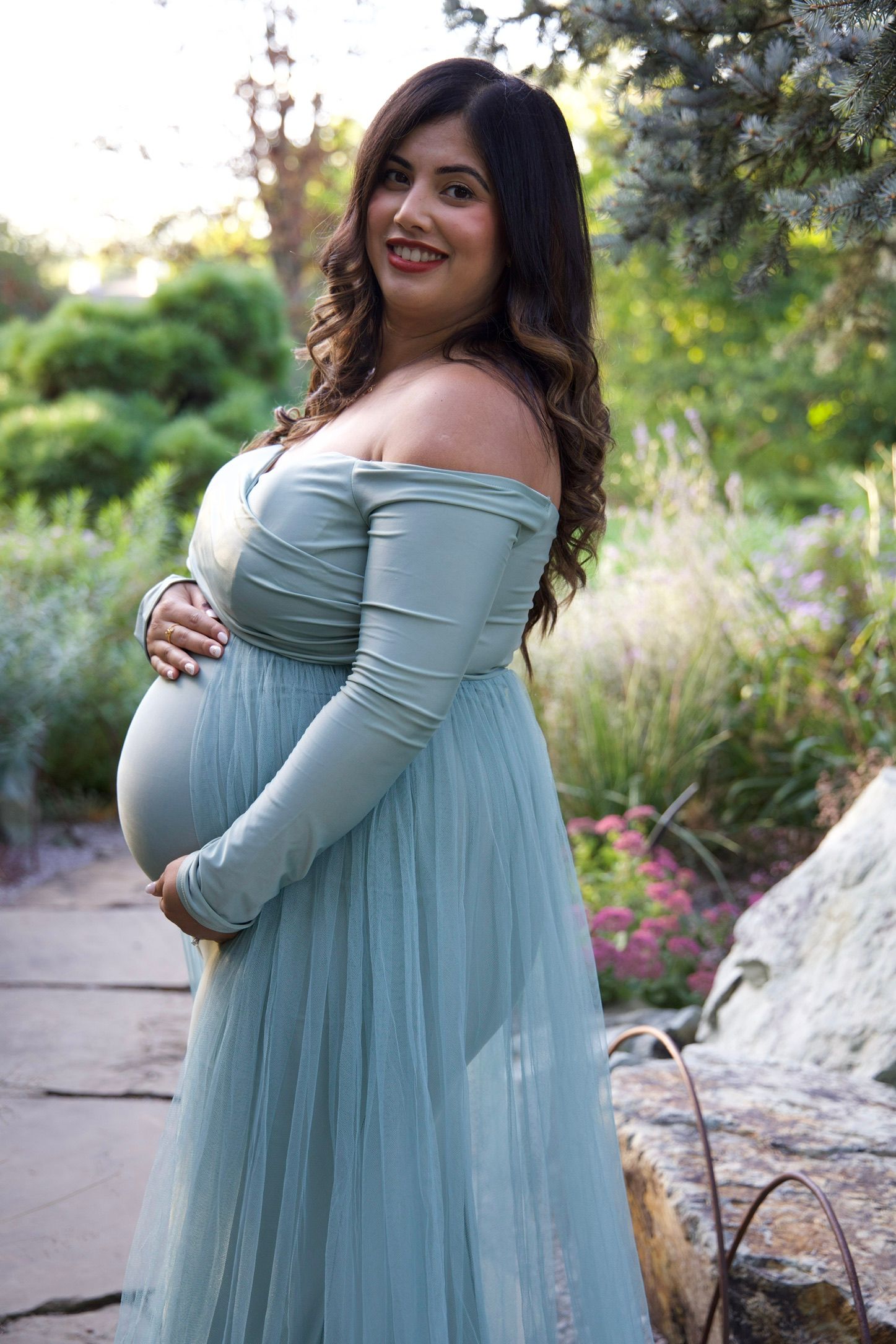 Glowing pregnant woman in sage green off-shoulder tulle gown cradling baby bump in a garden setting.