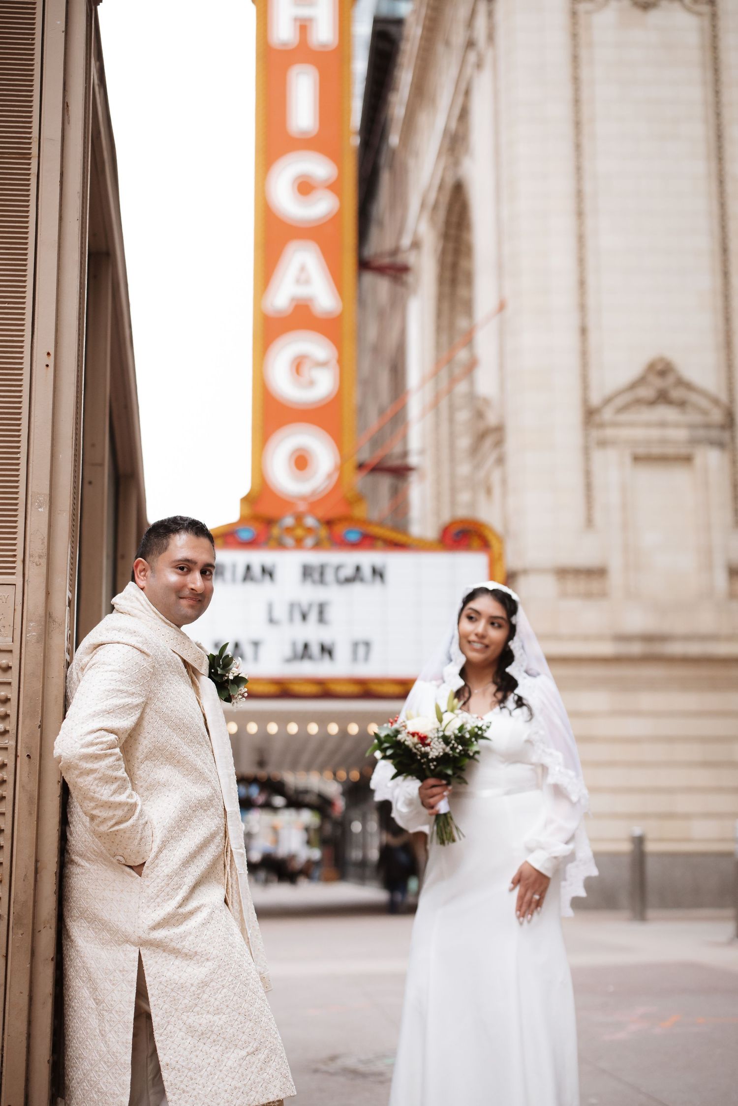 Groom leans against wall while bride holds bouquet near Chicago Theatre marquee sign.