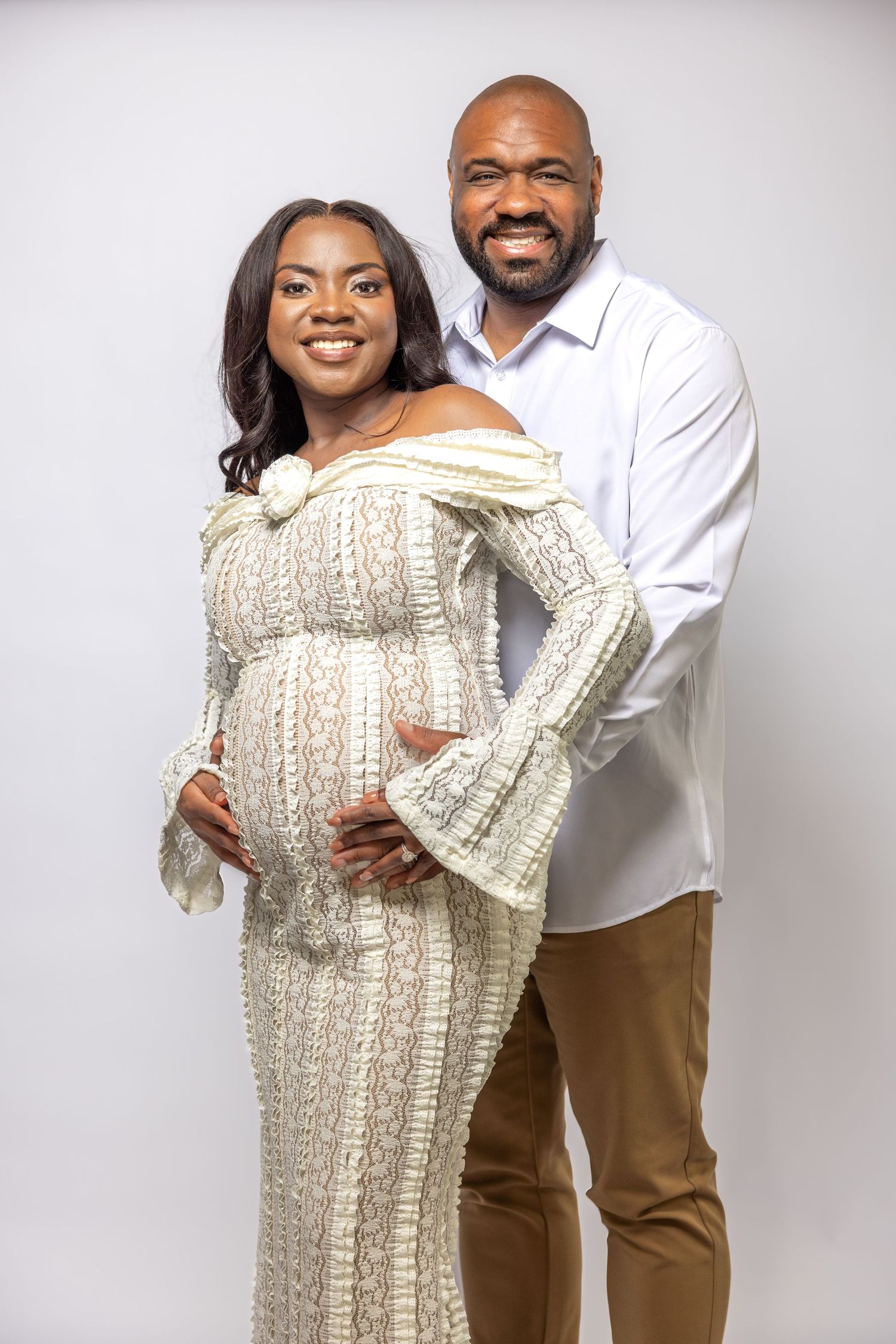 Smiling pregnant woman in off-shoulder cream knit gown posing with partner during maternity photoshoot.