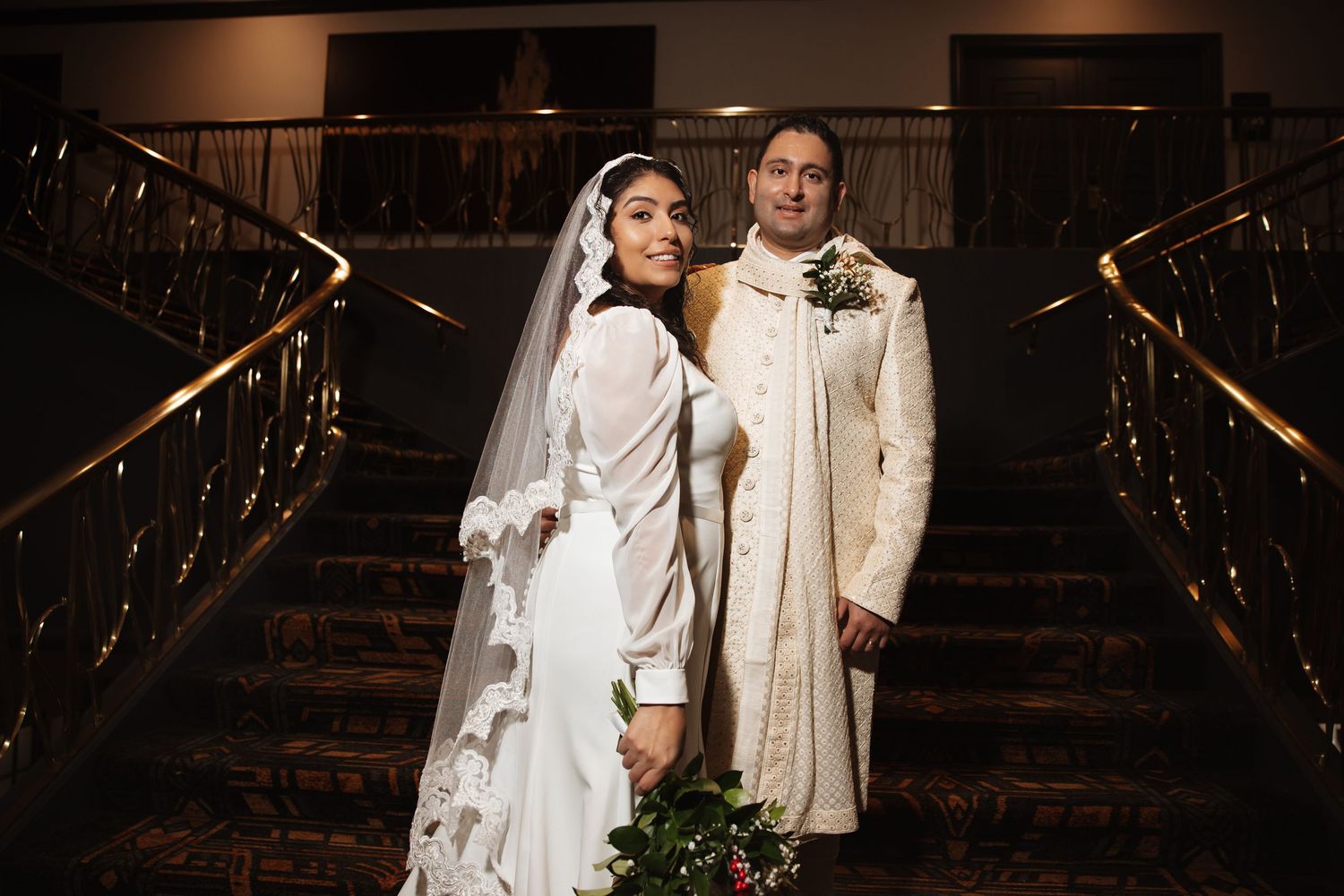 Bride holding bouquet and groom in sherwani pose on grand staircase with gold railings.