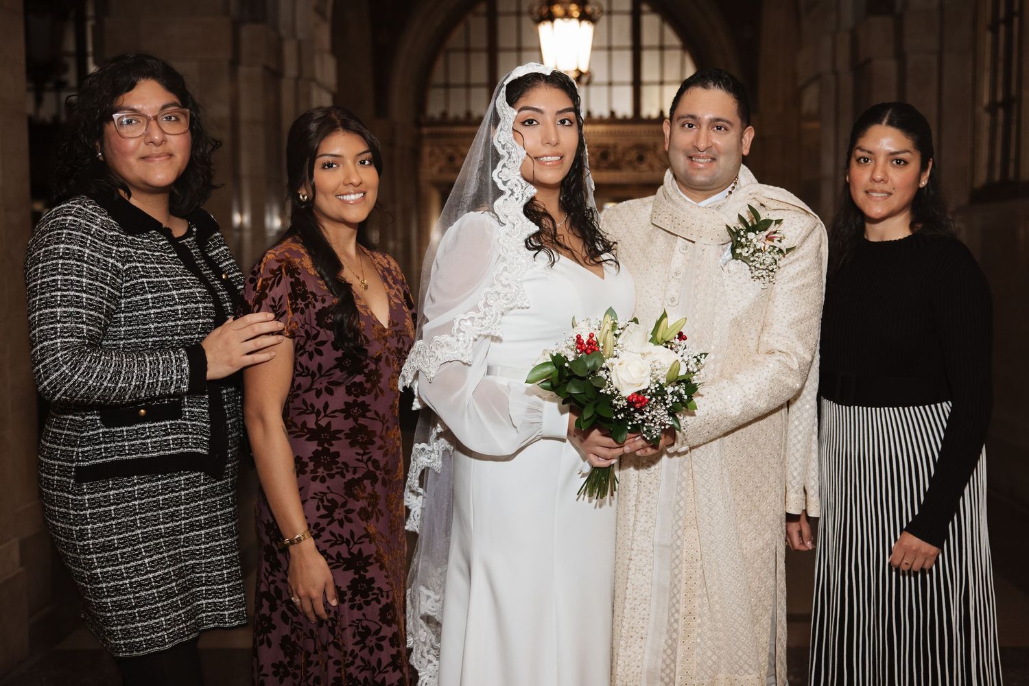 Bride in white gown and veil holds bouquet while posing with groom in ivory sherwani and three guests at wedding.