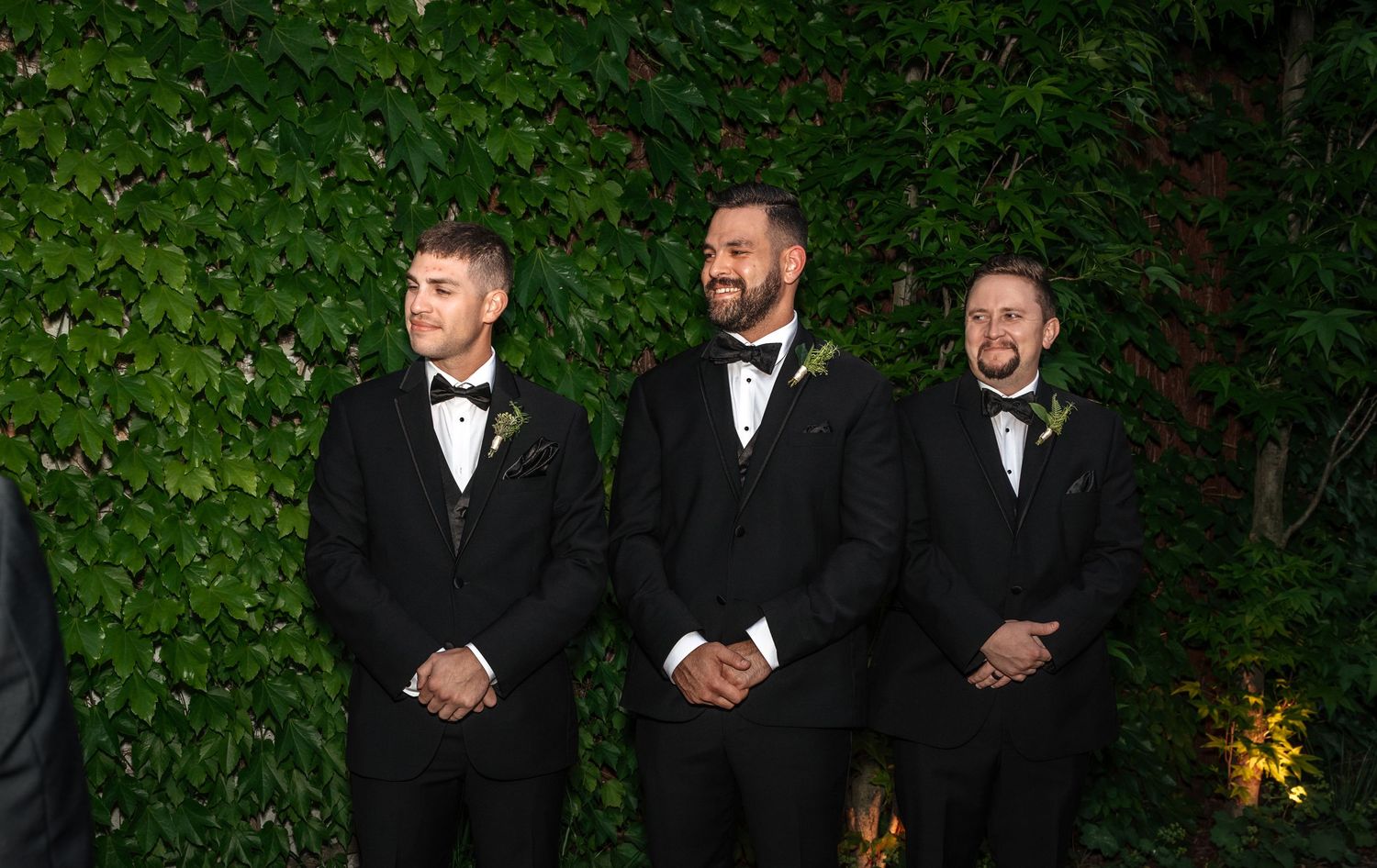 Three groomsmen in black tuxedos stand together smiling during an outdoor wedding ceremony.