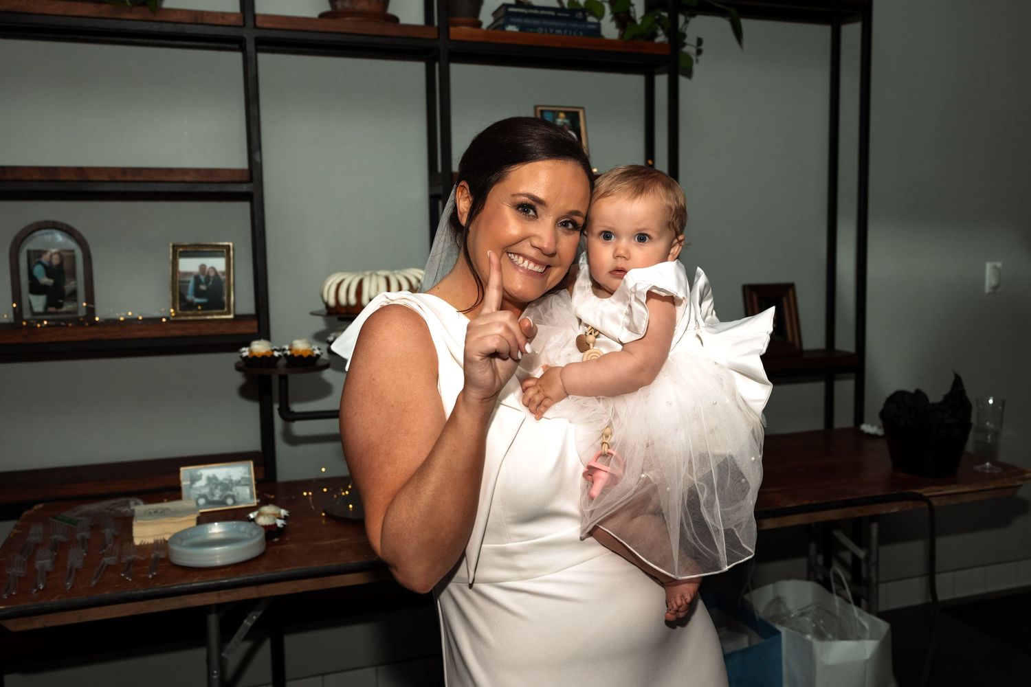 Bride in white wedding dress poses smiling while holding baby in white dress at wedding venue.