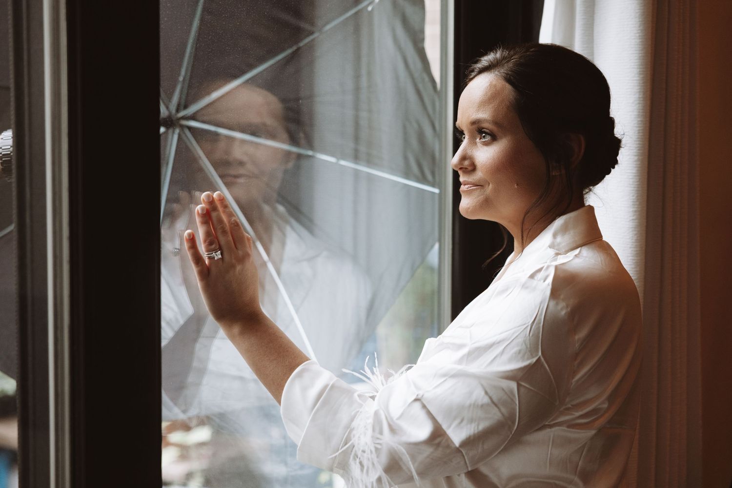 Beautiful bride in white robe touching window glass, soft natural light illuminating her face.