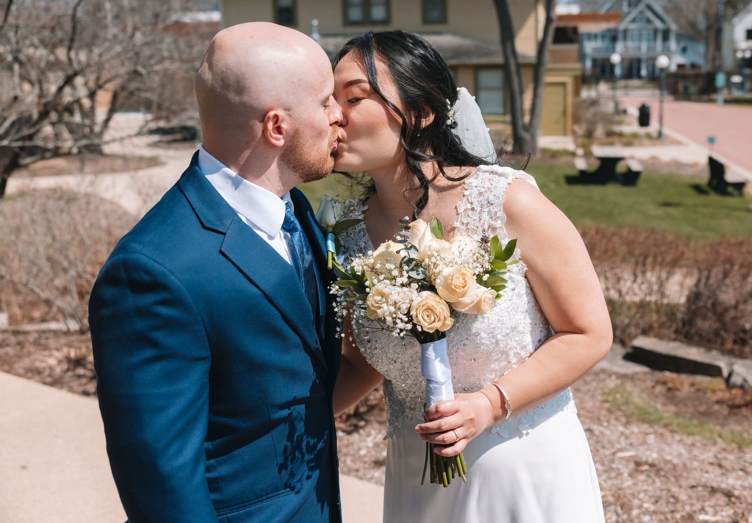 Bride and groom kiss outdoors on their wedding day, bride holding a beautiful bouquet of white and cream flowers.