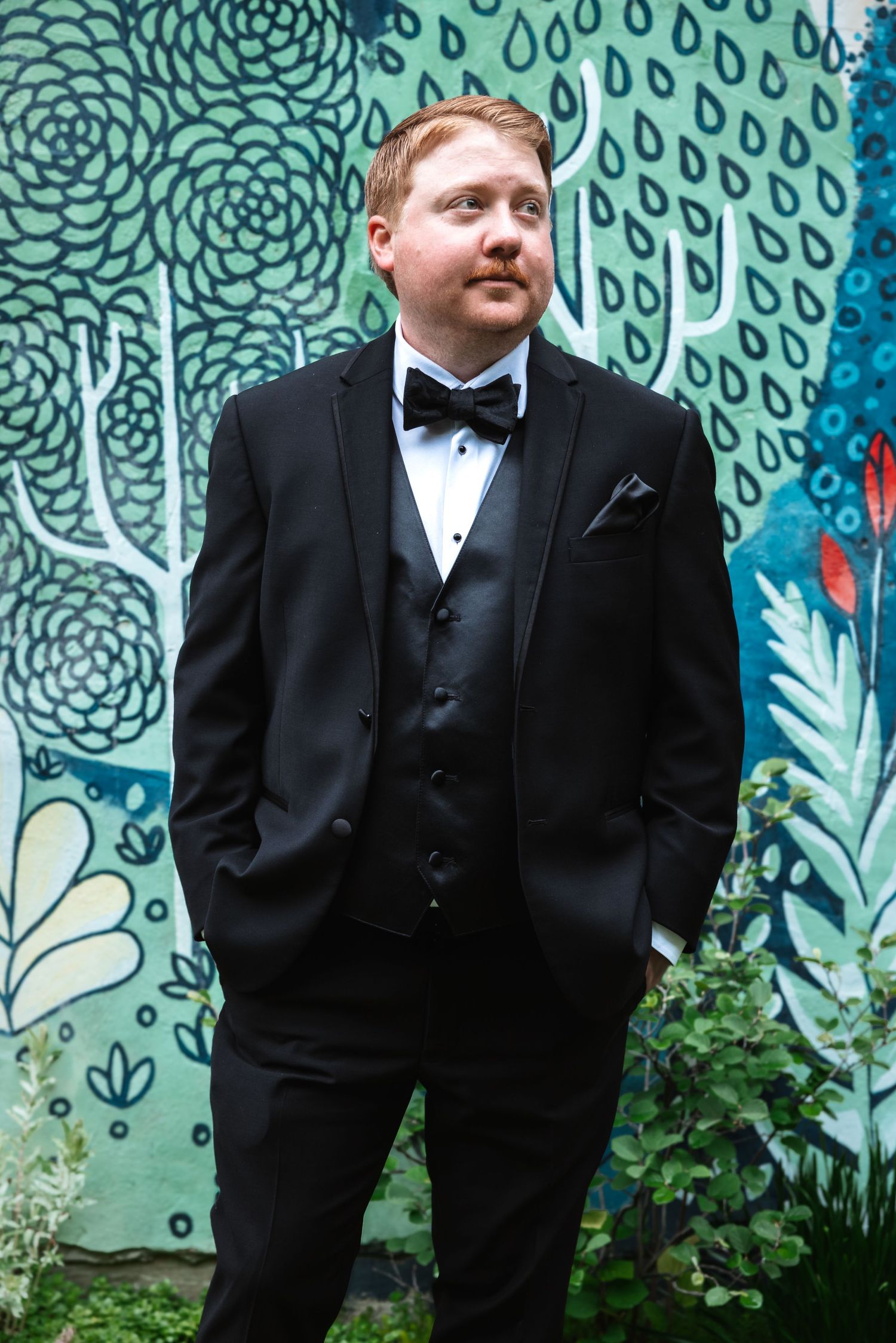 Dapper groom in black tuxedo and bow tie poses with hands in pockets before a colorful floral mural.