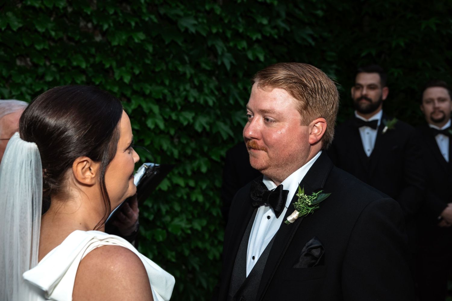 Groom in black tuxedo gazes at bride in white dress during outdoor wedding ceremony.