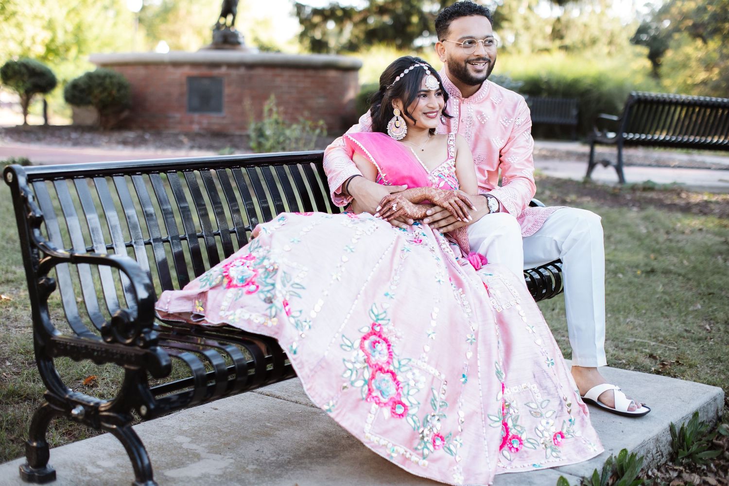Indian bride and groom sitting on a park bench in pink traditional wedding outfits.