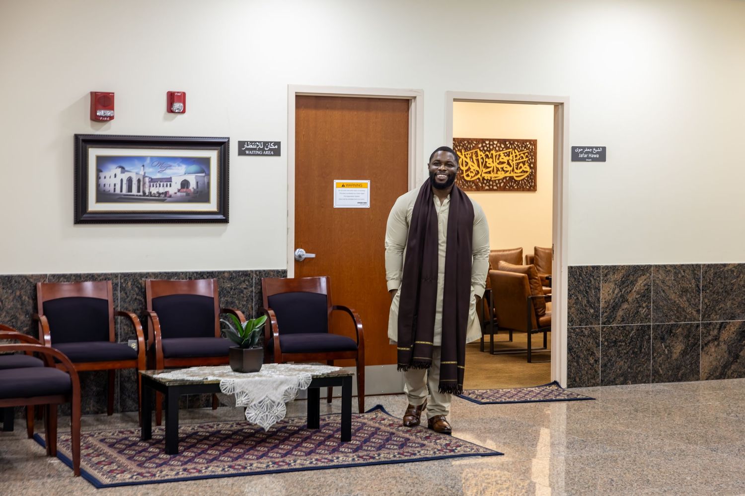 A man in a white outfit and brown scarf stands alone in a mosque lobby near chairs and a rug.