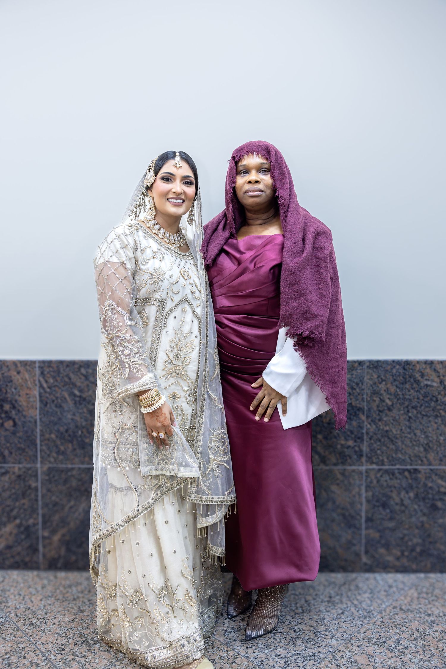 A South Asian bride in white embroidered attire smiles beside a woman wearing a burgundy dress and draped headscarf.