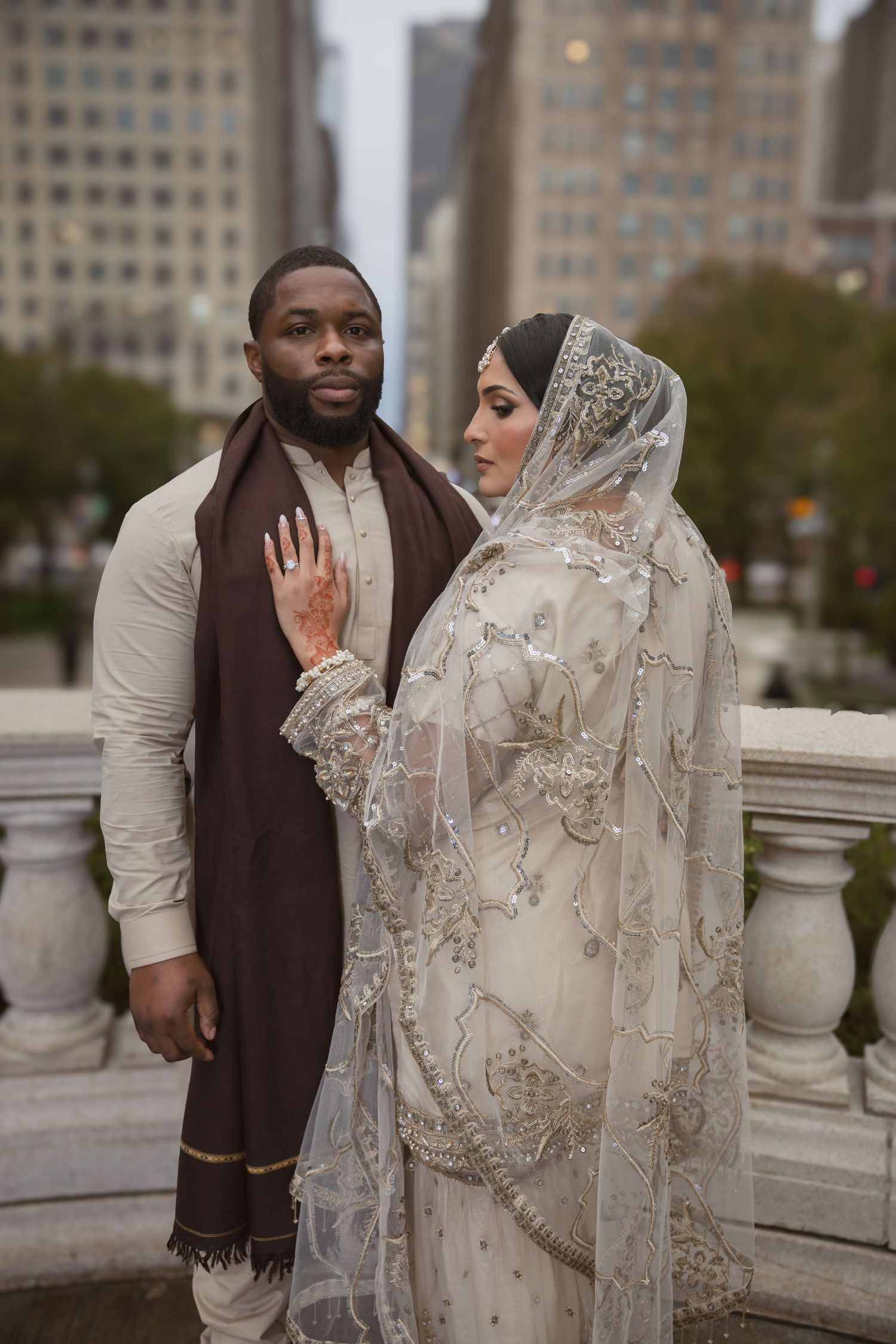 Interracial couple in traditional Islamic wedding attire pose outdoors with city skyline backdrop.