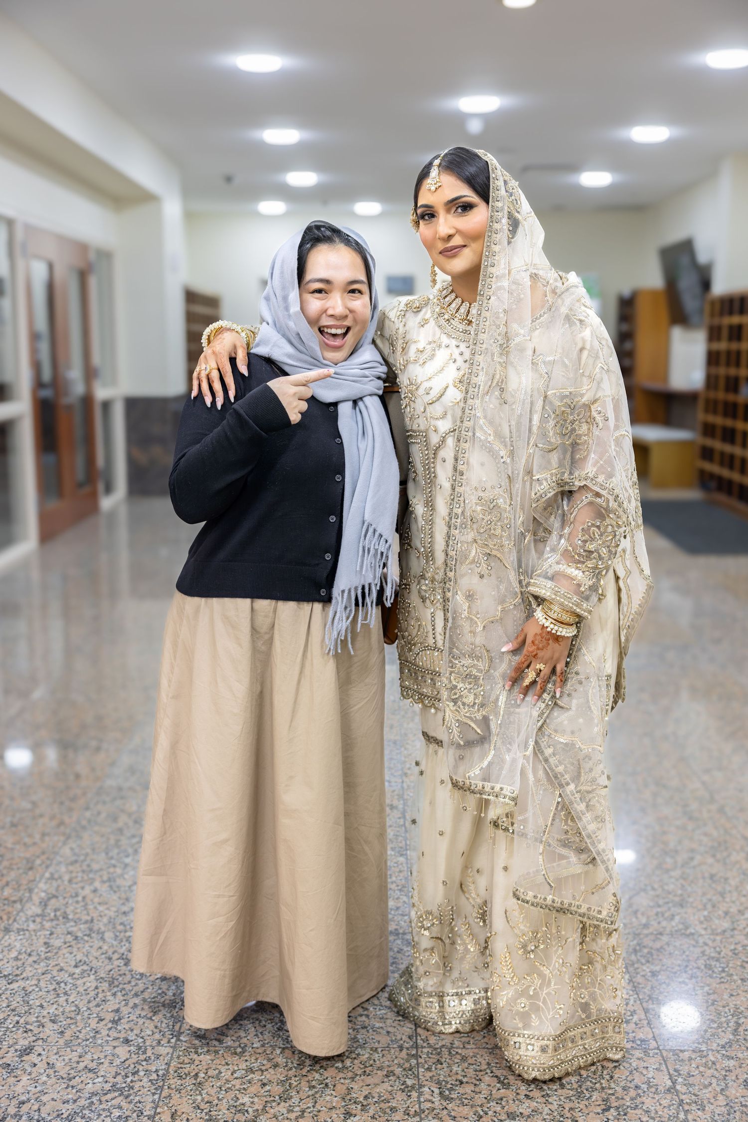Muslim bride in gold embroidered white lehenga poses with smiling woman in hijab at wedding venue.