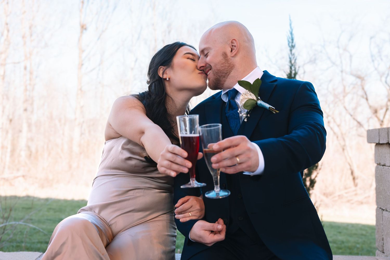 Couple sharing a romantic kiss while holding red champagne glasses at their outdoor wedding ceremony.