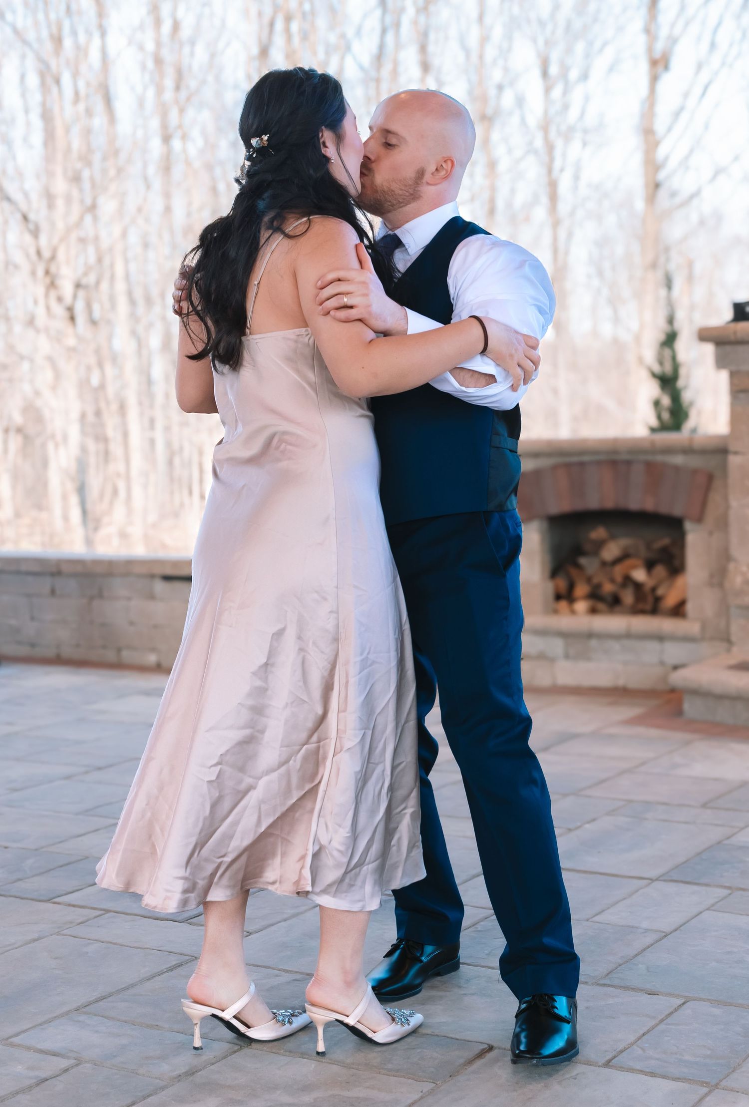 Bride in blush gown and groom in navy vest share a romantic kiss on a stone patio at wedding.