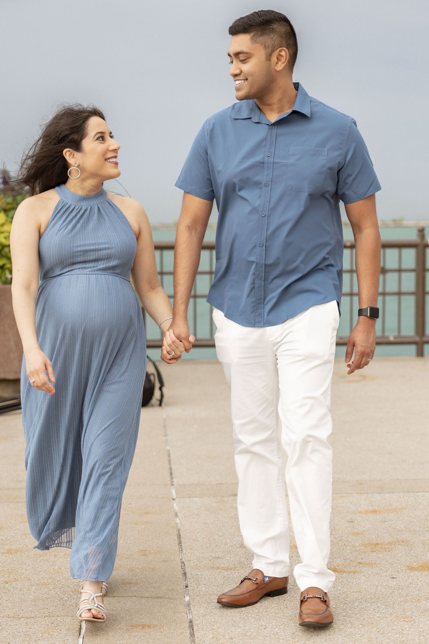 Smiling pregnant couple holding hands walking along waterfront promenade together.