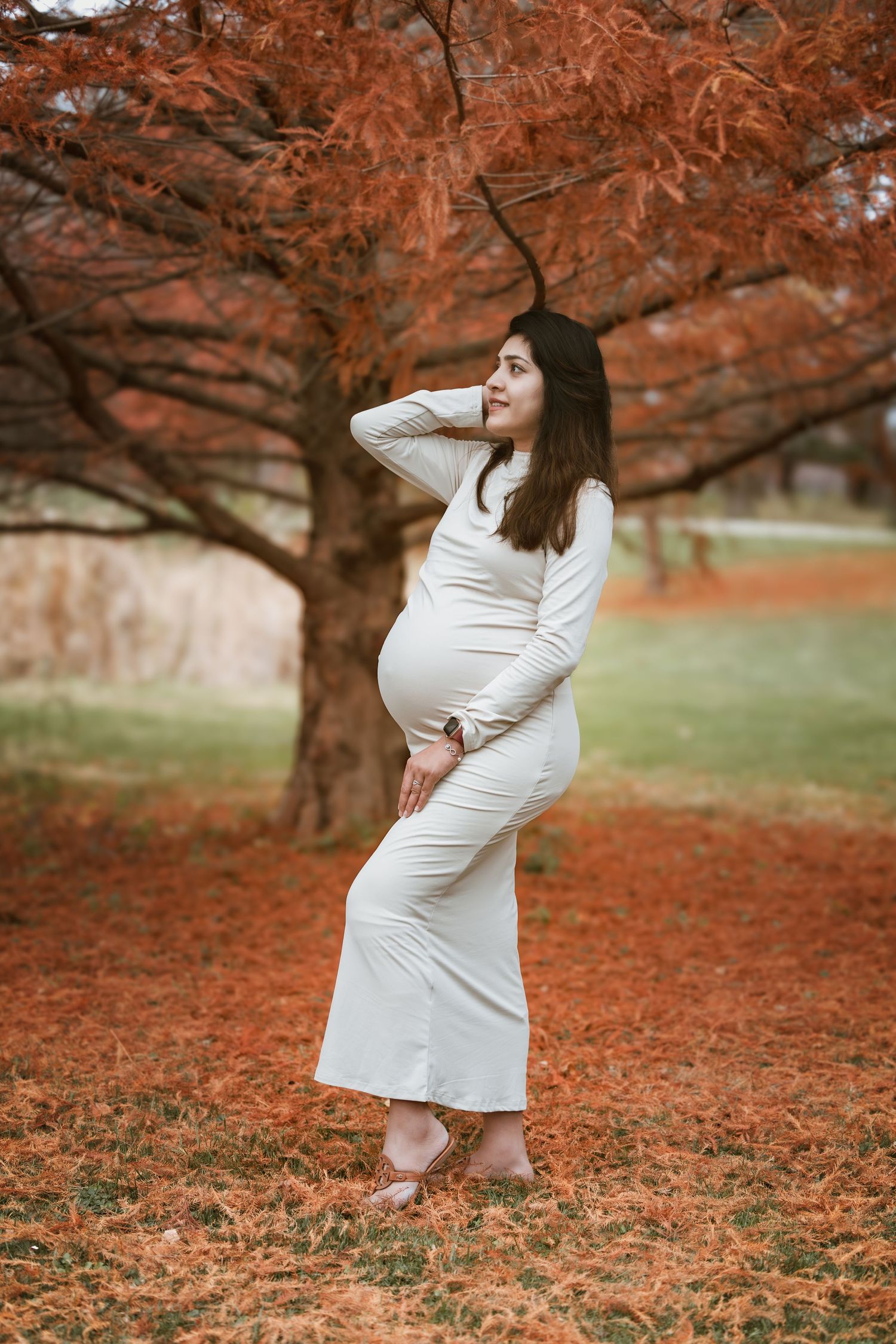 Pregnant woman in white dress posing under vibrant red autumn tree in park.