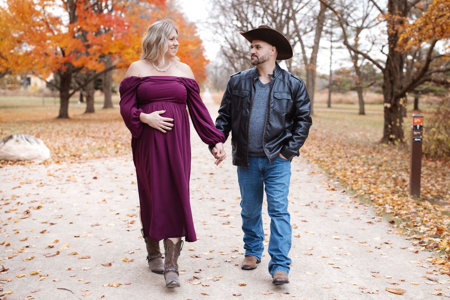 Glowing pregnant woman in burgundy dress walks hand-in-hand with cowboy partner in fall park.