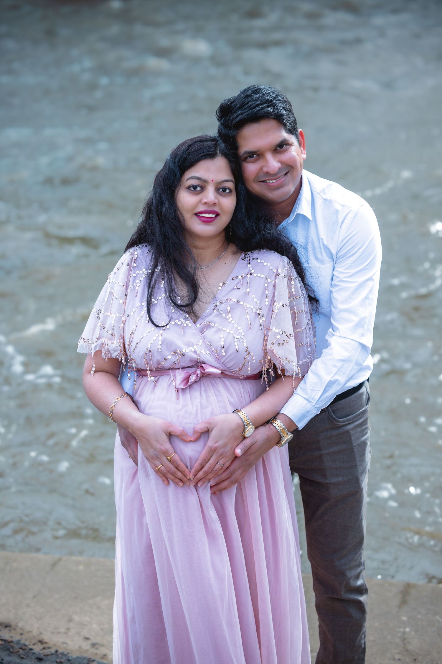 Indian couple sharing a romantic maternity portrait outdoors near water, both cradling the pregnant belly together.