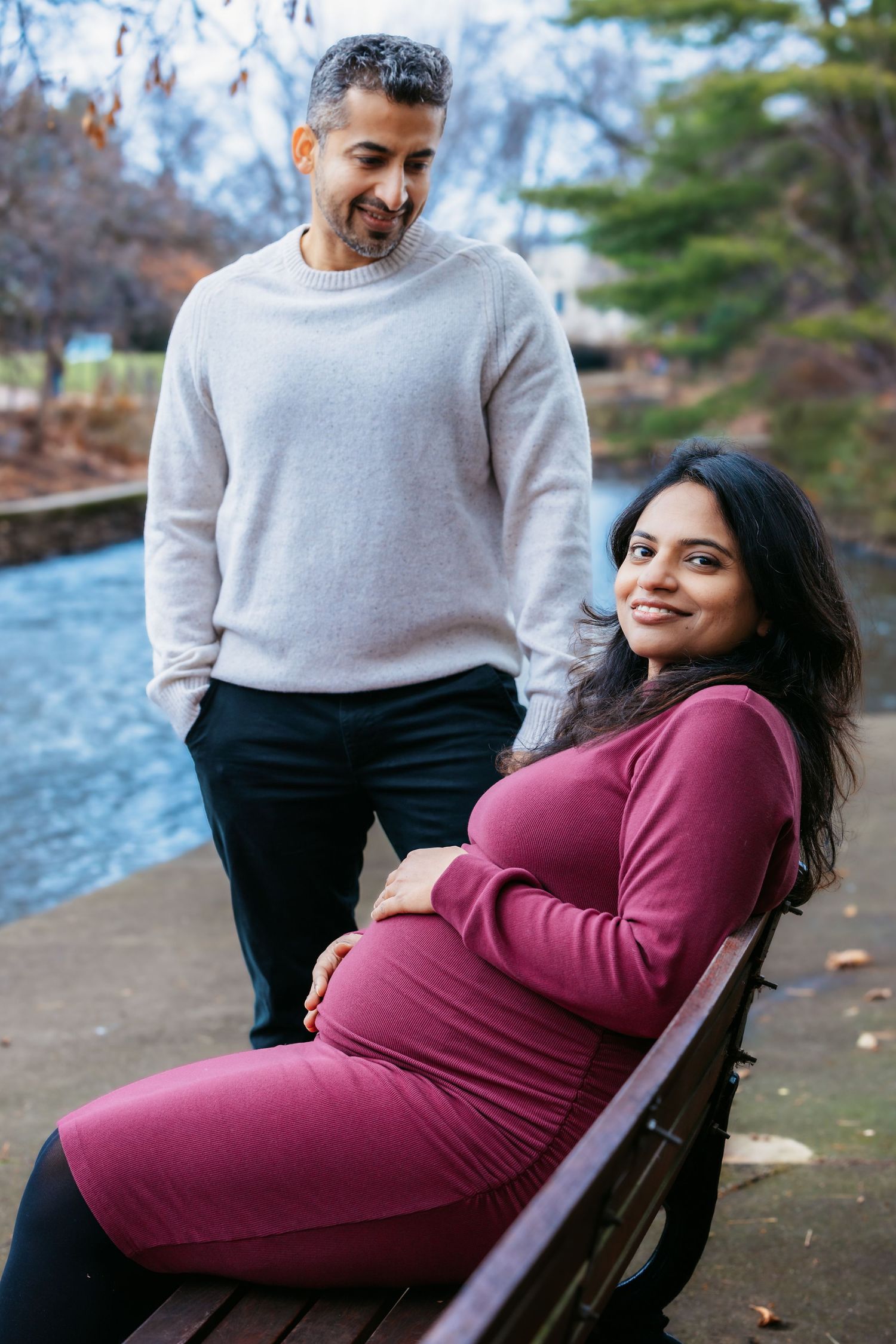 Pregnant woman in burgundy dress sits on bench as her partner stands beside her near a park pond.