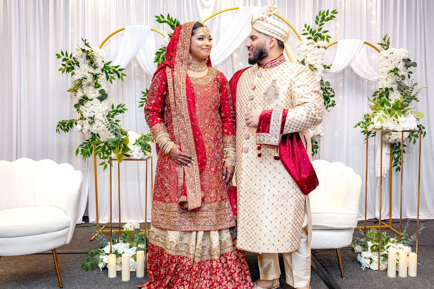 South Asian bride and groom in traditional red and cream wedding outfits posing before white drape backdrop.