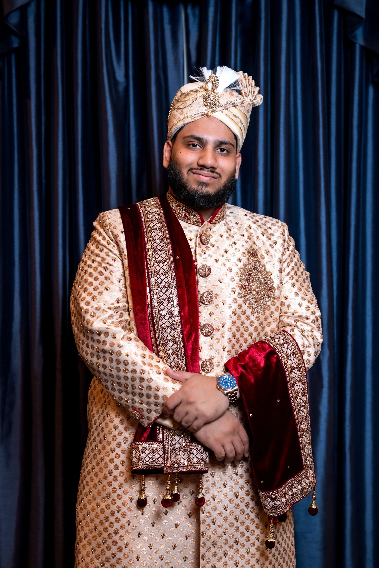 Smiling Indian groom wearing gold embroidered sherwani and decorative turban posing before blue curtains.